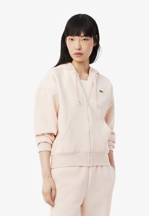 Light pink zip-up hoodie made from soft fabric, featuring a kangaroo pocket, drawstring hood, and ribbed cuffs. Lacoste logo on chest.