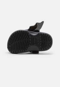 Černá gumová sandála s texturovanou podrážkou, která má hluboké drážky a zvýšený střed pro lepší trakci. Zakřivený detail popruhu na zadní straně.