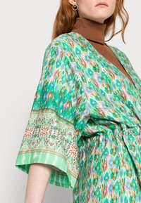 Vestido verde com padrão, apresentando um decote em V, mangas largas e uma silhueta fluida. Detalhes notáveis incluem uma cintura decorativa e realces.