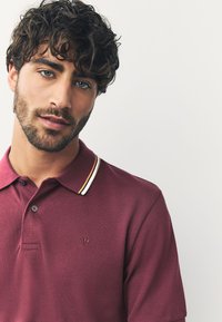 Bordeauxrotes Poloshirt mit strukturierter Oberfläche, ausgestattet mit einem kontrastierenden Kragen mit gelben und weißen Streifen sowie einem kleinen, aufgestickten Kronenlogo auf der Brust.
