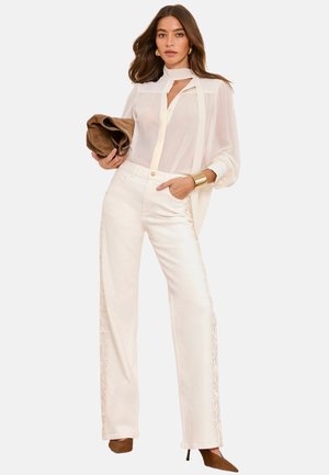 Woman wearing sheer white blouse with scarf detail, high-waist wide-leg white pants with side embroidery, brown pointed heels, holding a brown clutch.