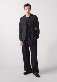 Veste de costume noire ajustée avec des revers crantés, associée à un t-shirt noir à col rond et un pantalon noir à jambes larges. Des chaussures noires brillantes complètent le look.