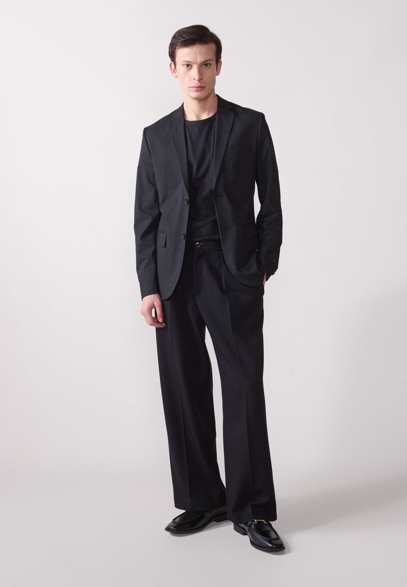 Sacou negru croit, cu revere normale, asociat cu un tricou negru cu gât rotund și pantaloni negri cu picioare largi. Pantofii negri lucioși completează ținuta.