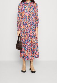 Kukallinen midi-mekko, jossa on kirkkaan oransseja, vaaleanpunaisia ja vihreitä kuvioita sinisellä taustalla, puffeilla pitkillä hihoilla ja löysällä istuvuudella. Ruskea käsilaukku ja beige nilkkurit täydentävät asua.