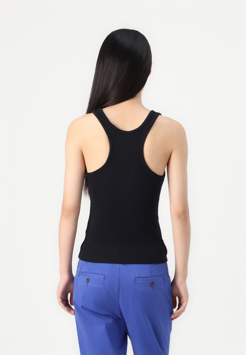 Schwarzes Tanktop mit schmalen Trägern, figurbetontem Design und glatter Textur. Kombiniert mit hochsitztenden blauen Hosen mit Gesäßtaschen.