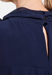 Blusa azul marino con cuello drapeado, cierre de botones en la parte trasera y un brillo sutil, fabricada en un tejido suave y ligero.