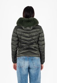 Giacca imbottita verde oliva con cappuccio peloso, motivo trapuntato, design aderente e polsini elastici, abbinata a jeans in denim azzurri.