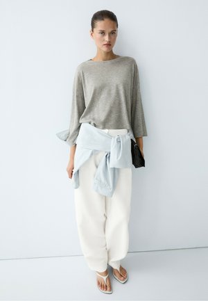 Donna che indossa una maglietta grigia a maniche lunghe, pantaloni bianchi, camicia azzurro chiaro annodata in vita, sandali infradito bianchi e tiene una borsa nera.