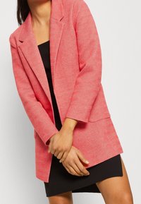 Blazer rosa a spina di pesce in tessuto testurizzato con rever a notched e apertura frontale. Indossato sopra un vestito nero, che mette in mostra una vestibilità rilassata.
