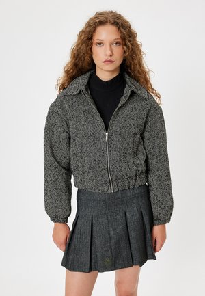 Houndstooth-mønstret sort og hvid cropped jakke med lynlås, elastisk kant og ribbet krave, parret med en plisseret grå nederdel.