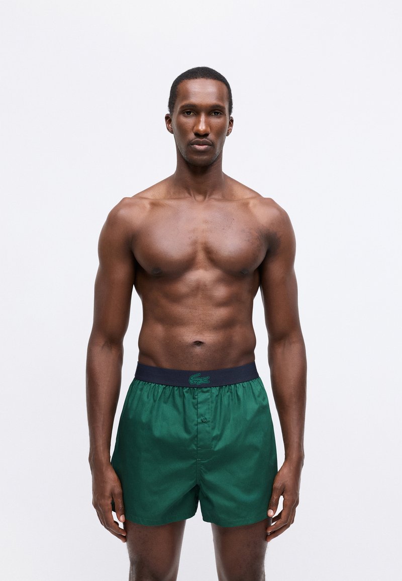 Green men's swim shorts made of smooth fabric, featuring a dark navy elastic waistband with a subtle logo. Simple design, regular fit.