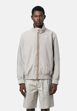 Uomo che indossa una giacca con cerniera grigio chiaro a collo alto e pantaloncini abbinati fino al ginocchio, in piedi davanti a uno sfondo semplice.