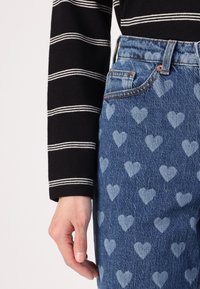 Jeans iz denima z lahkimi modrimi srčastimi vzorci, zaključeni s klasičnim dizajnom s petimi žepi in rjavim šivom, v kombinaciji z črno črtasto majico.