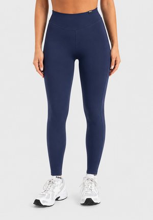 Femme portant un legging bleu marine taille haute et des chaussures de sport blanches, debout devant un fond clair uni.