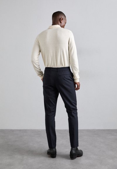 Cămașă polo lungă, de culoare bej deschis, ajustată, asortată cu pantaloni închiși navy, având talie elastică și buzunare la spate, purtată cu pantofi negri.