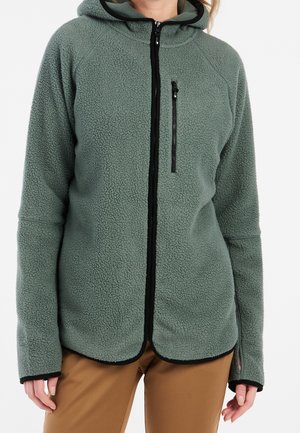 Personne portant une veste polaire zippée verte avec une poche zippée poitrine et des trous pour les pouces, associée à un pantalon marron.