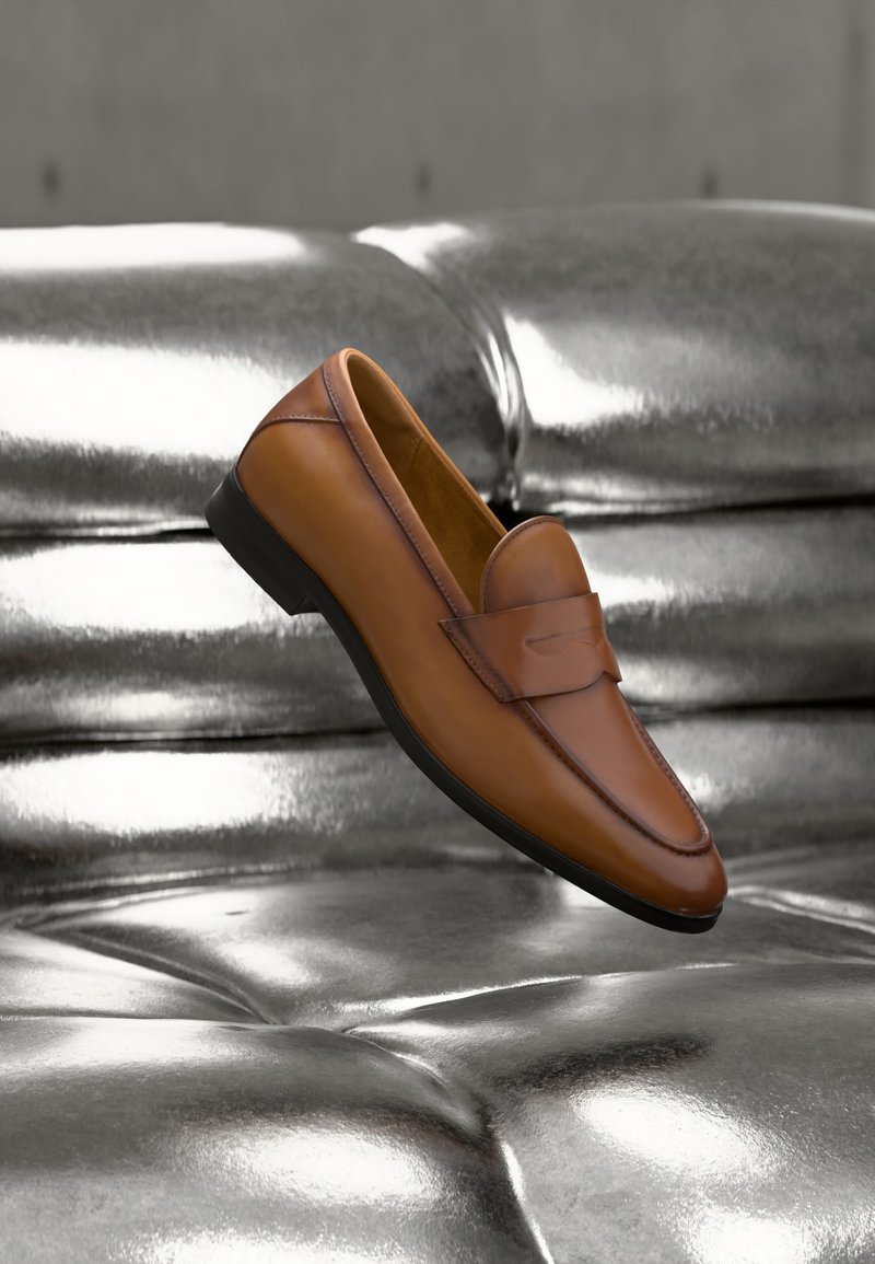 Mocassin en cuir marron flottant au-dessus d'un canapé en cuir argenté brillant et capitonné.