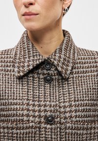 Brauns houndstooth mētelis ar augstu apkakli, ar melniem pogām un teksturētu vilnas maisījuma audumu. Tuvplāns fokuss uz apkakli un pogām.