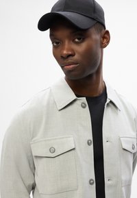 Ανοιχτό γκρι πουκάμισο με δύο τσέπες στο στήθος, υφή υφάσματος και ένα μαύρο καπέλο. Ένα σκούρο μπλουζάκι από κάτω είναι μερικώς ορατό.