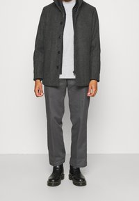 Grau melierter Wollmix-Mantel mit durchgehender Knopfleiste und Kapuze, kombiniert mit einem hellgrauen Hemd. Dunkelgraue maßgeschneiderte Hose und schwarze Stiefel vervollständigen den Look.