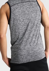 Camiseta atlética sin mangas de color gris con ribetes negros, llevada por una persona de pie vista desde atrás, con los brazos relajados a los lados.