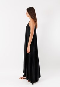 Abito lungo nero con un design fluido, realizzato in tessuto leggero. Presenta spalline sottili e uno spacco laterale, abbinato a sandali marroni.