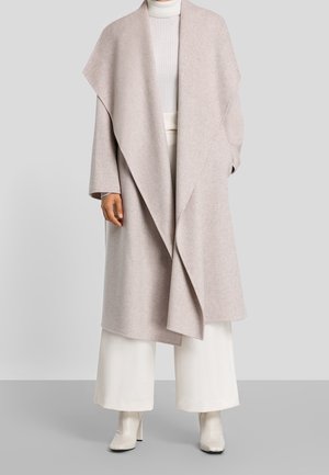 Woman wearing a light beige oversized wool coat, white wide-leg pants, white turtleneck, and white heeled ankle boots, standing on a gray floor.