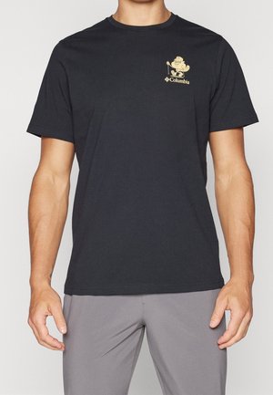 Mann trägt ein schwarzes kurzärmliges T-Shirt mit einem beigen Wandercartoon und dem "Columbia"-Logo auf der linken Brust, kombiniert mit grauer Hose.