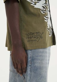 Olijfgroen shirt met wit grafisch ontwerp, korte mouwen, een lossere pasvorm en geborduurde branding "DON:ED HARDY designs" aan de zijkant.