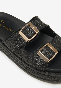 Sandali con piattaforma neri a trama intrecciata, dotati di doppie fibbie metalliche. La suola è texturizzata e leggermente rialzata, offrendo supporto.