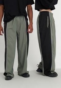 Athletic pants in olive green and black, featuring white side stripes, elastic waist, and zippered pockets. Smooth, lightweight fabric.