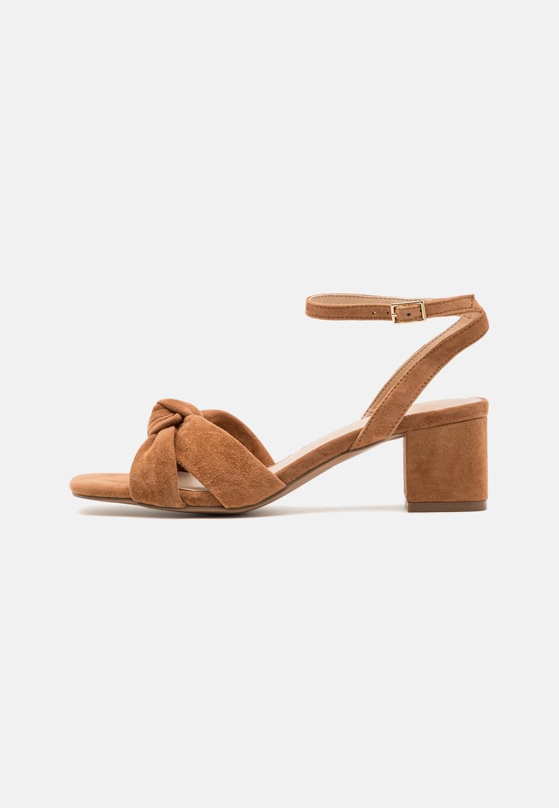 Sandales à talons en daim marron avec un design noué à l'avant, une bride à la cheville, et un talon bloc. Caractérisées par une texture lisse et une structure minimaliste.