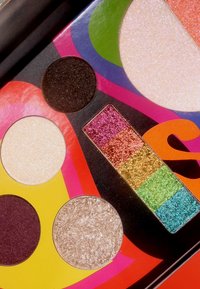 Fargerik øyenskyggepalett med fem sirkulære panner: svart, gull, beige og to skimrende nyanser, pluss en rektangulær glitrende stripe i regnbuens farger.