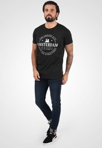 Sort t-shirt med hvid Amsterdam grafik, korte ærmer, rund halsudskæring. Kombineret med mørkeblå jeans og sorte sneakers med gyldne detaljer.