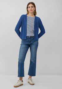 Vrouw in een blauwe cardigan, gestreept wit-blauw shirt, cropped blauwe spijkerbroek en witte sneakers, staande tegen een effen grijze achtergrond.