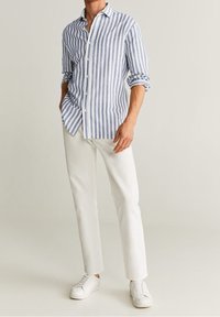 Muž ve modro-bílém pruhovaném dlouhém rukávu s vyhrnutými rukávy, bílých kalhotách a bílých teniskách na jednotném pozadí.