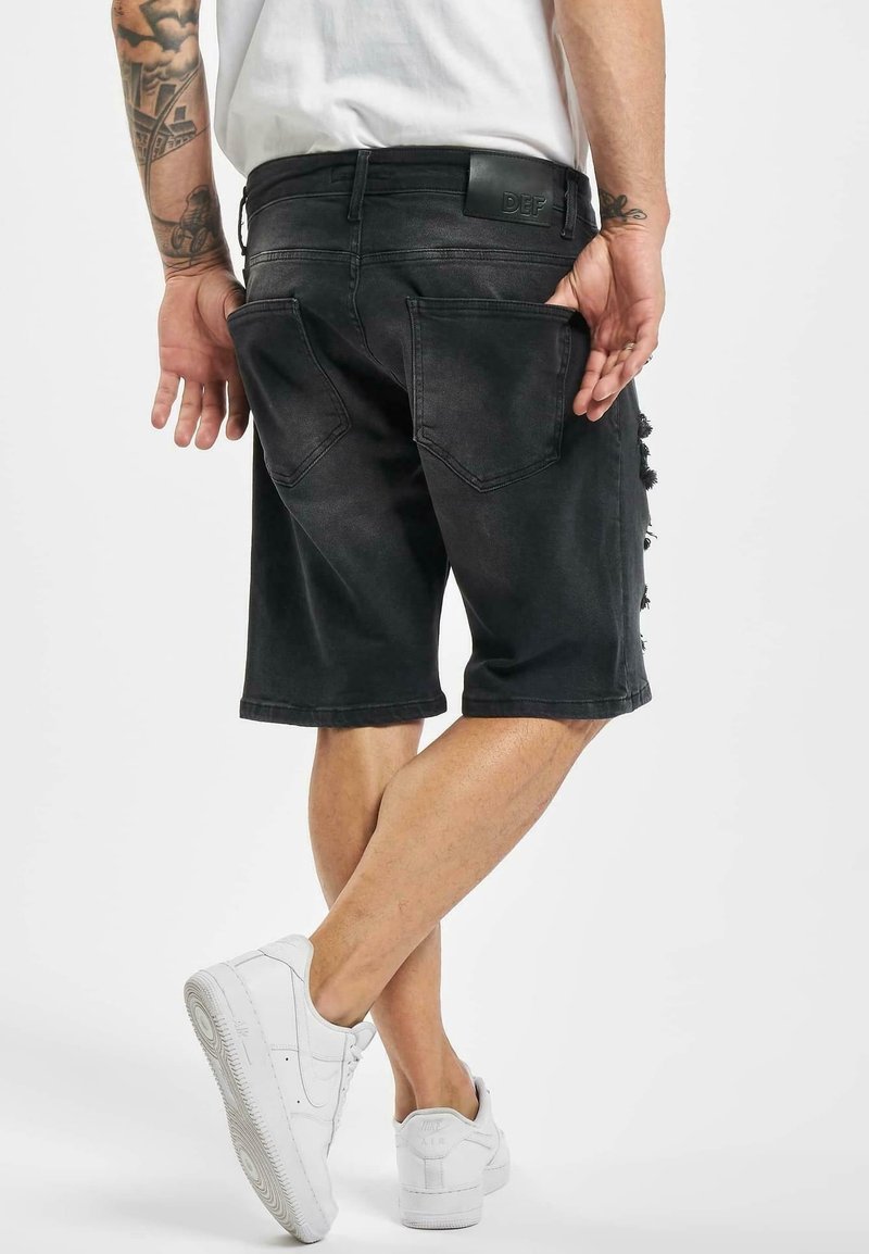 Homme portant un short en jean noir et des baskets blanches, avec les bras tatoués, debout, jambes croisées et mains près des poches arrière.