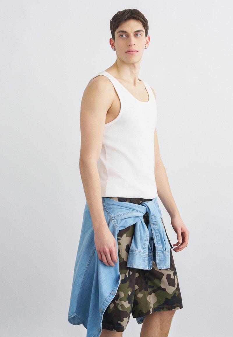 Giovane uomo con canottiera bianca, pantaloncini mimetici, giacca di jeans blu legata in vita, in piedi contro uno sfondo chiaro uniforme, guarda verso sinistra.