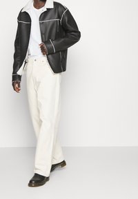 Uomo che indossa una giacca in shearling nera, una t-shirt bianca, pantaloni bianchi larghi e stivali in pelle nera, in piedi contro uno sfondo chiaro e semplice.