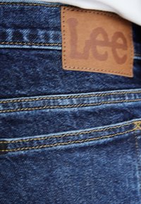 Denim jeans in dark blue with tan leather logo patch labeled "Lee." Features gold stitching and a classic five-pocket design.