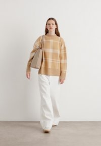 Sweater beige xadrez com gola alta, combinado com calças brancas de perna larga. O modelo carrega uma bolsa bege clara. Texturas suaves predominam.