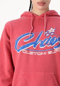 Rød hettegenser med en frontlomme, med et blått grafisk design og teksten "Chaos Custom Supply." Laget av bomull med en myk tekstur.