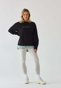 Czarny sweatshirt z napisem "Pilates Princess", nałożony na jasnoniebieską koszulę, w zestawie z jasnoszarymi legginsami i butami sportowymi.