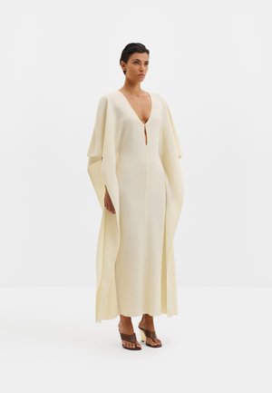 Femme portant une longue robe fluide couleur crème avec des manches larges et un décolleté en V profond, assortie de sandales à talons marron, debout sur un fond blanc.