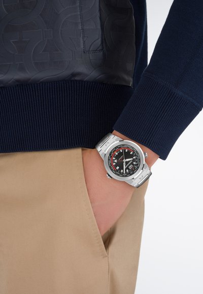 Persona que lleva un reloj de pulsera de metal plateado con una esfera detallada en negro y rojo, suéter azul marino y pantalones beige con la mano en el bolsillo.