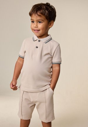 Ung dreng med kort brunt hår iført beige polotrøje og matchende shorts, står med den ene hånd i lommen og smiler let.