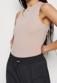 Vaaleanpunainen hihaton toppi korkealla pääntiellä, jossa on valkoinen Nike-logo rinnassa, yhdistettynä joustaviin mustiin housuihin, joissa on naruvyötärö.