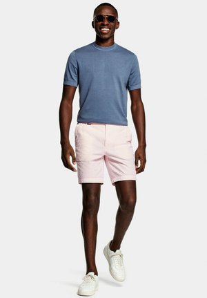 Jonge man met een zonnebril met ronde glazen, een blauw overhemd met korte mouwen, roze shorts en witte sneakers, lopend met een glimlach op een witte achtergrond.