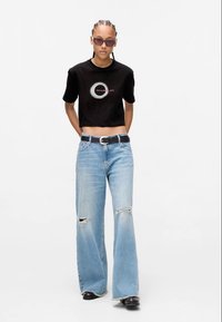 Camiseta preta cropped com gráfico circular, combinada com jeans largos rasgados em azul claro. Os acessórios incluem um cinto preto e óculos de sol.
