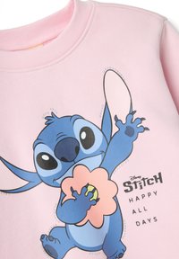 Sweat-shirt rose avec un motif de Stitch tenant une fleur, accompagné d'un texte noir indiquant "HAPPY ALL DAYS" et des accents décoratifs.